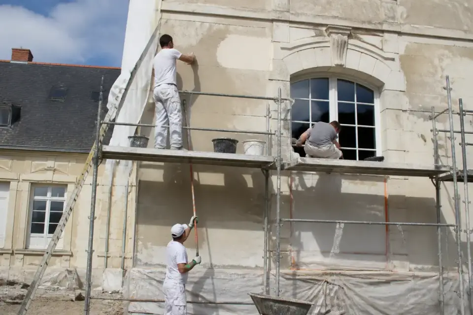 Trois ouvriers restaurent la façade d'un bâtiment sur échafaudage, appliquant enduit et peinture murale.