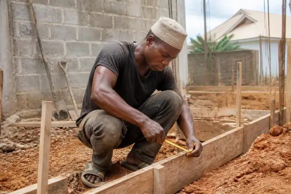 Ouvrier mesurant des planches de bois sur un chantier de construction en plein air, portant une casquette.