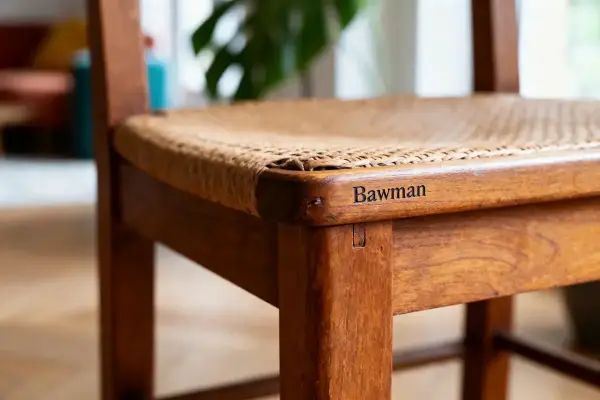 Chaise en bois avec assise tissée, gravée "Bawman" sur la structure. Ambiance intérieure floue en arrière-plan.