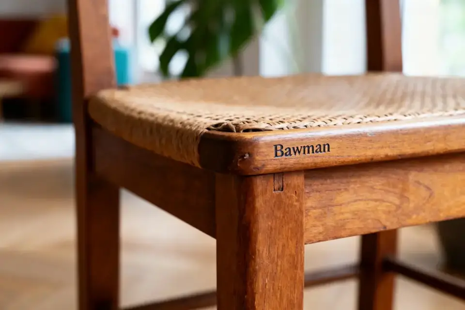 Chaise en bois avec assise tissée, gravée "Bawman" sur la structure. Ambiance intérieure floue en arrière-plan.