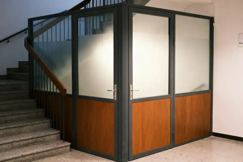 Cabine individuelle en bois et verre sous des escaliers, avec une porte grise à poignée métallique.