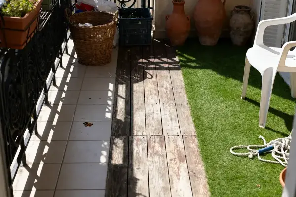 Balcon avec carreaux, bois, gazon artificiel, chaise blanche, paniers, pots de terre cuite, corde et rai de lumière.