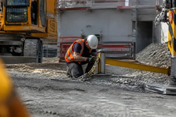 Travailleur de chantier, casque et gilet, manœuvre un équipement près d'une pelleteuse sur terrain en construction.