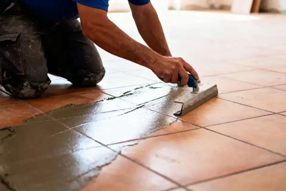 Personne appliquant du mortier sur carrelage à l'aide d'une taloche, genoux à terre, vêtement de travail.