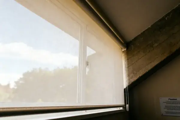 Store enrouleur beige couvrant une fenêtre, ciel bleu partiellement visible à travers le tissu blanc.